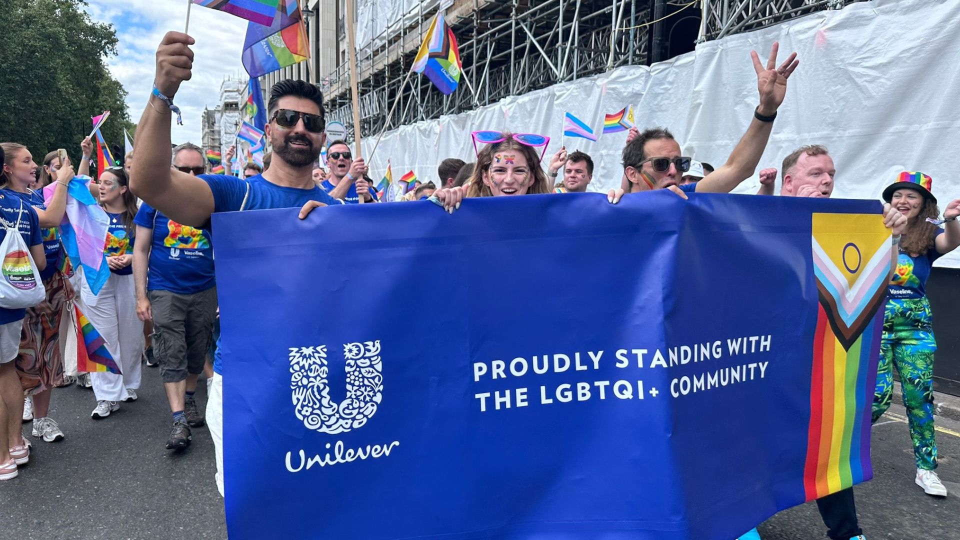 A group of people from Unilever walking and waving flags at Pride London, whilst the people at the front hold a Unilever sign with text saying 'Proudly Standing with the LGBTQI+ Community'