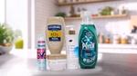 Five household and personal care products displayed on a kitchen counter: a bottle of Hellmann’s Real Mayonnaise, Degree Whole Body Deodorant spray, Dove Intensive Repair cream jar, Dove Pro-Ceramide body serum with pump, and Persil Wonder Wash laundry detergent in a green bottle. A blurred kitchen background with plants and utensils is visible.