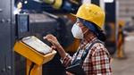 Woman factory worker in protective helmet, mask and coveralls operating a machine via a control panel.