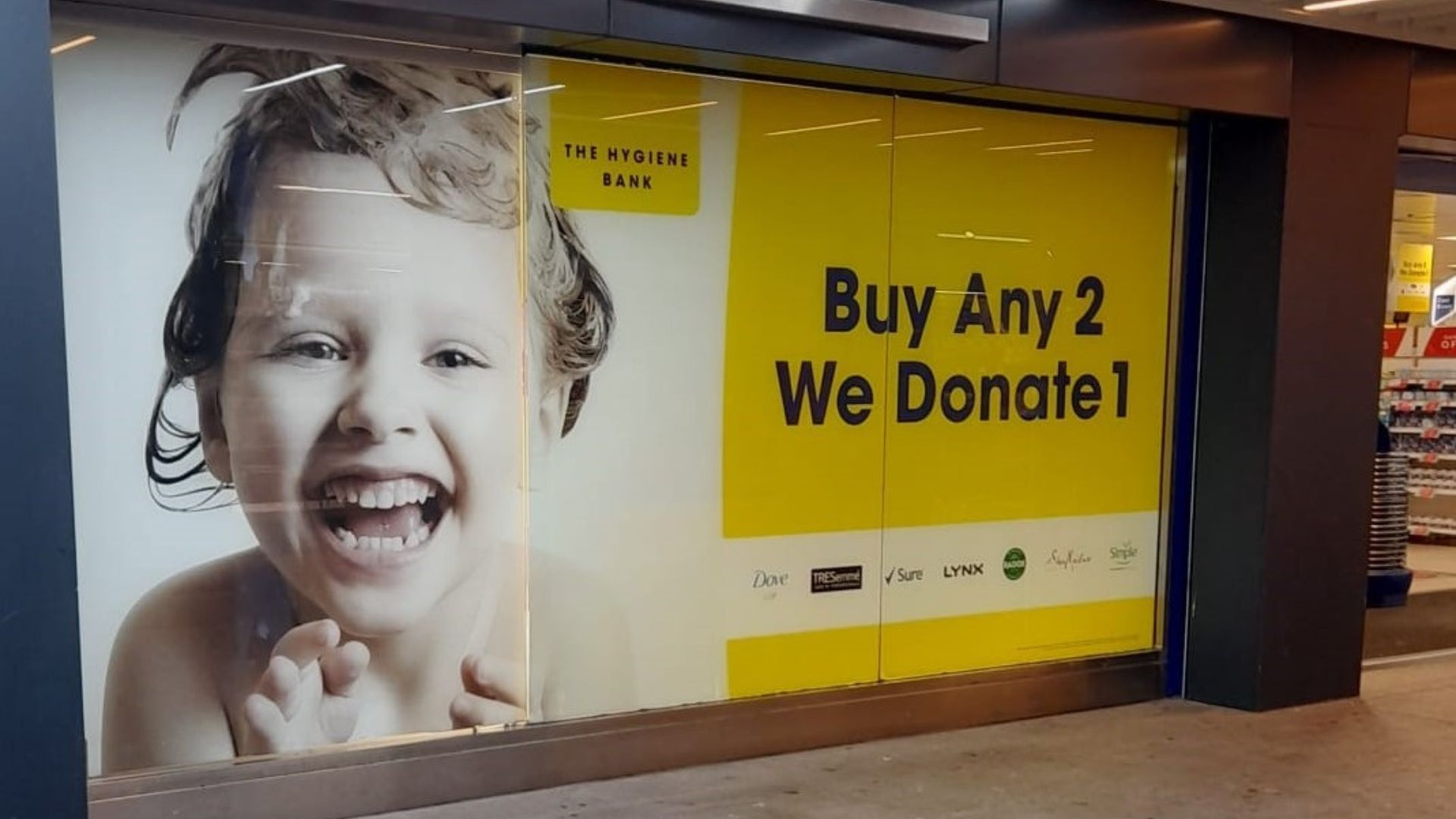 Large window sticker with text 'Buy Any 2 We Donate 1' against a yellow background, seven brand logos, and a photo of a child