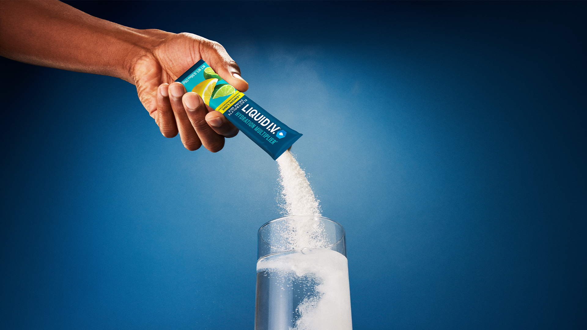 A hand pouring Liquid I.V. Hydration Multiplier powder from a sachet into a glass of water, with the powder visibly mixing against a blue background.