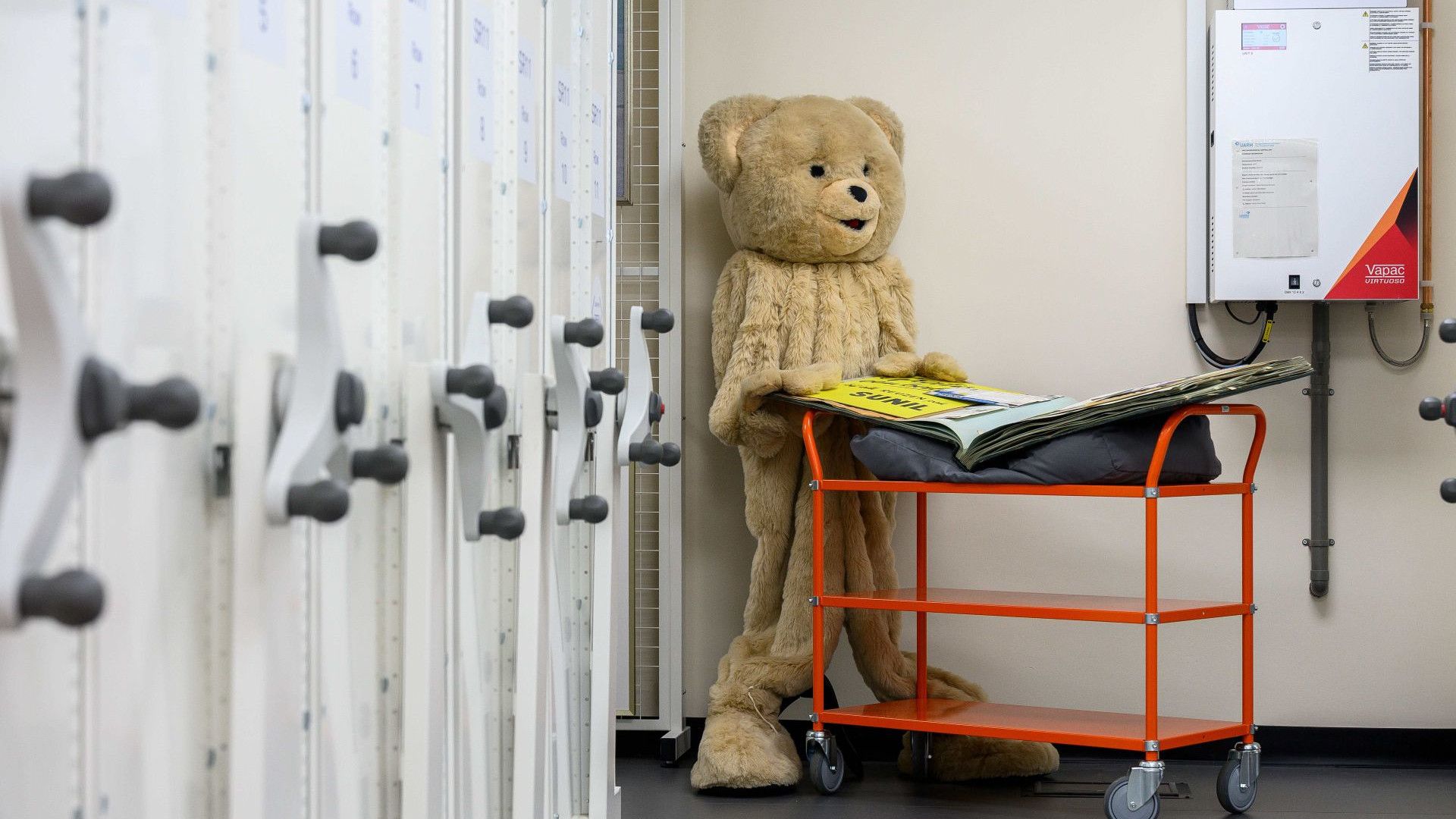 Large teddy bear stands beside an orange cart in an industrial storage room.