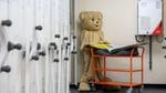 Large teddy bear stands beside an orange cart in an industrial storage room.