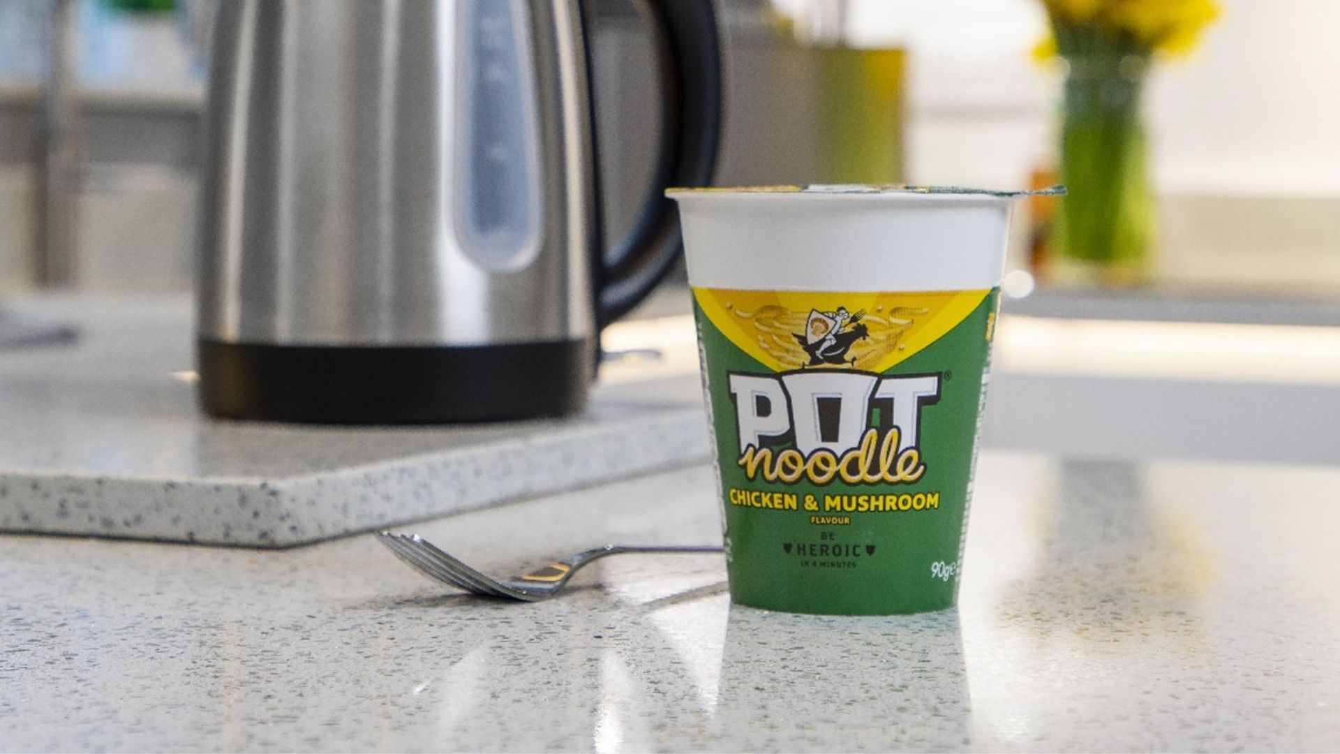 A Chicken & Mushroom Pot Noodle sitting on a grey kitchen counter, with a fork and kettle.