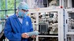 A person wearing a blue Unilever lab coat, hairnet, safety glasses and face mask examines a grey plastic bottle in a factory setting, with industrial equipment in the background.