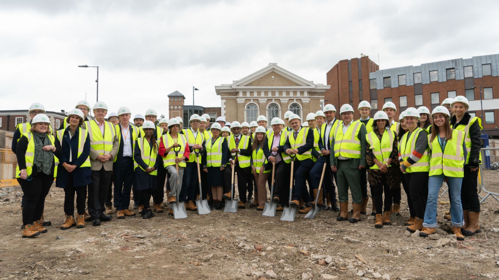 group of people wearing high-vis jackets and hardhats smiling at camera, six people holding spades