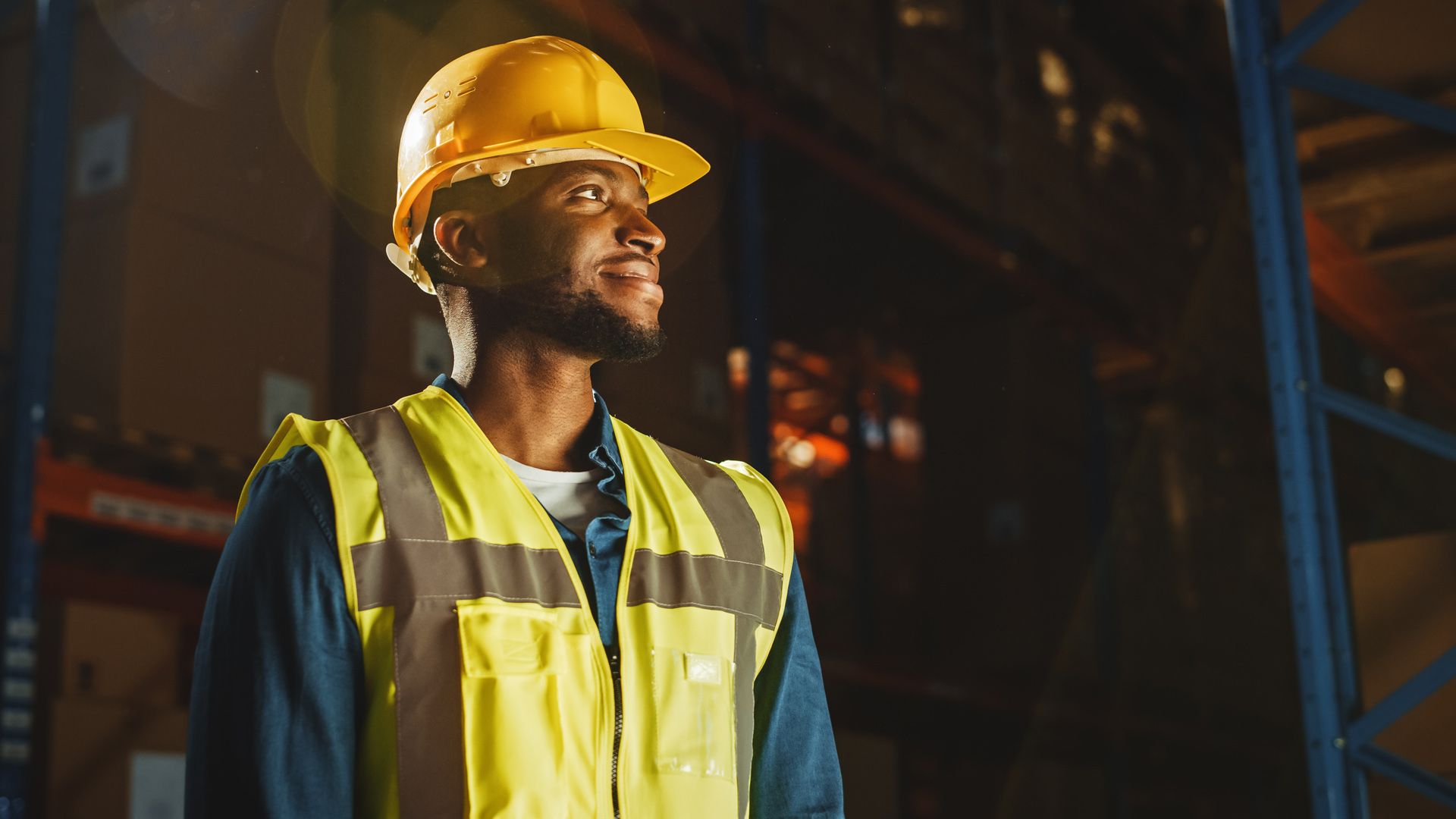 Male warehouse worker wearing safety vest and hard hat. In the background are shelves full of delivery goods.