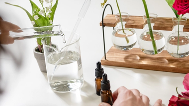 Image of three rounded test tubes in a wooden holder, containing a variety of flowers including a red rose. A technician is pouring liquid from a test tube into a glass bottle.