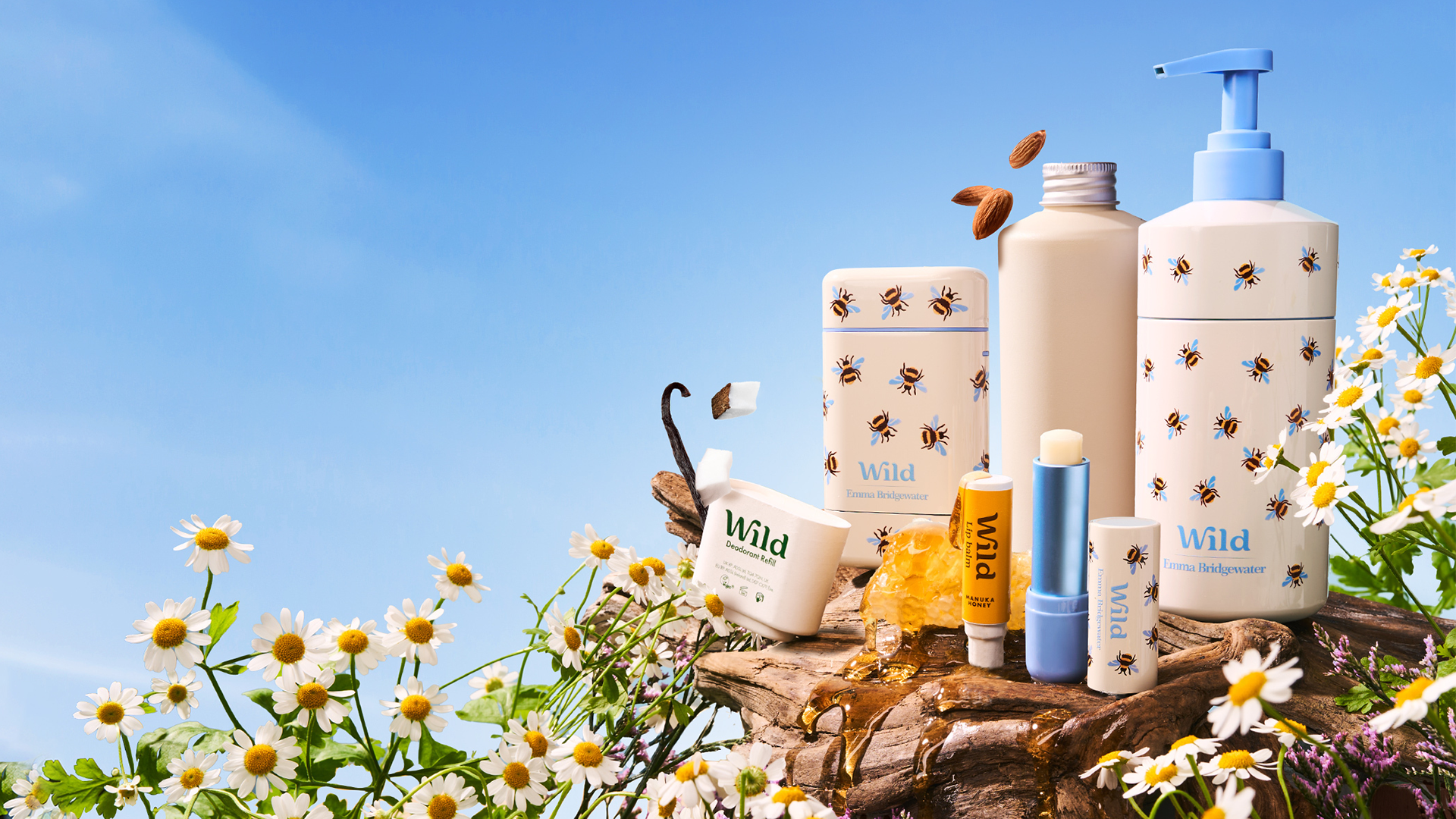 A selection from the Wild product range – a bottle, pump bottle, lip balm and deo stick – arranged on a lump of wood among white and yellow flowers.